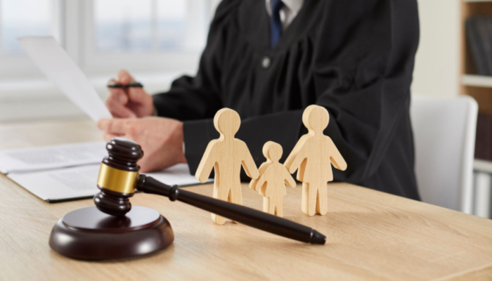 Gavel and little wooden figures of family up close on table in courthouse, and judge reading divorce settlement in background